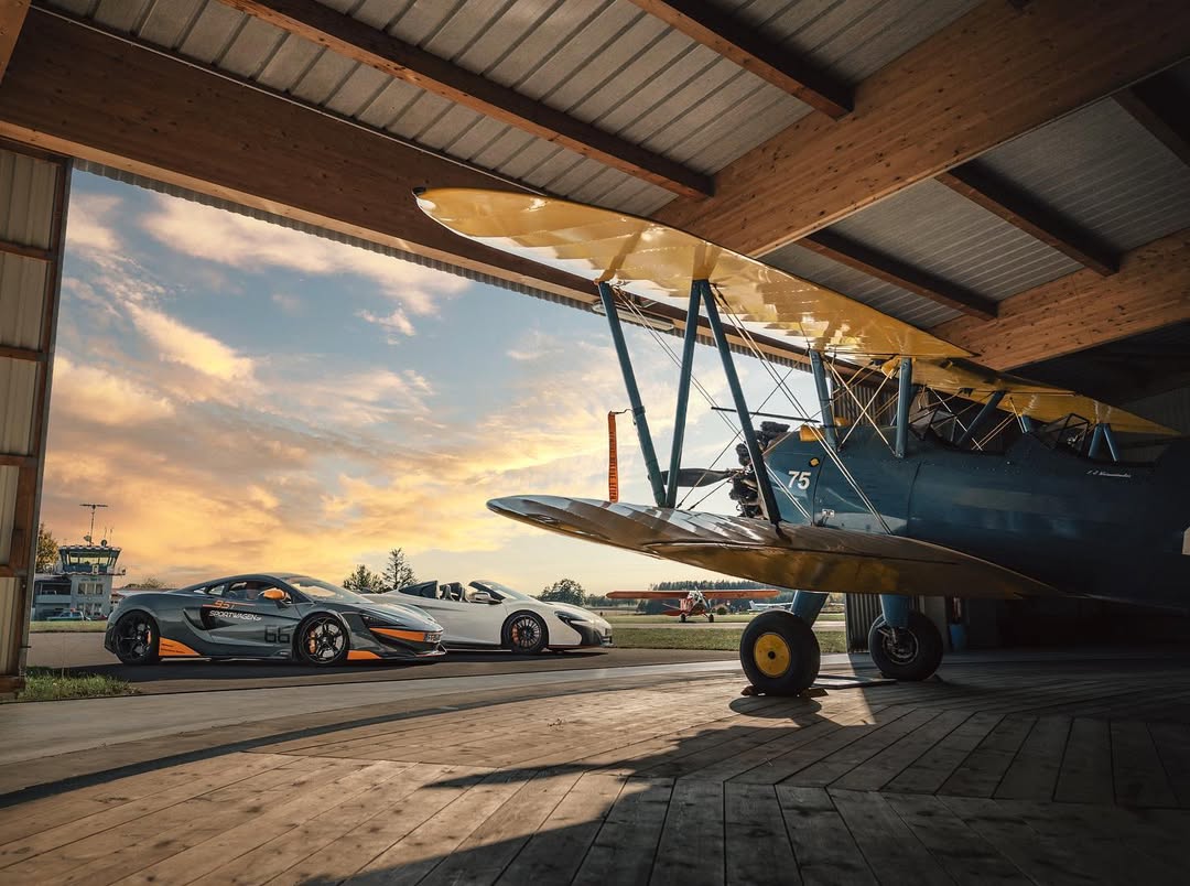 Ein historisches Flugzeug steht in einer Halle, während moderne Sportwagen davor parken.
